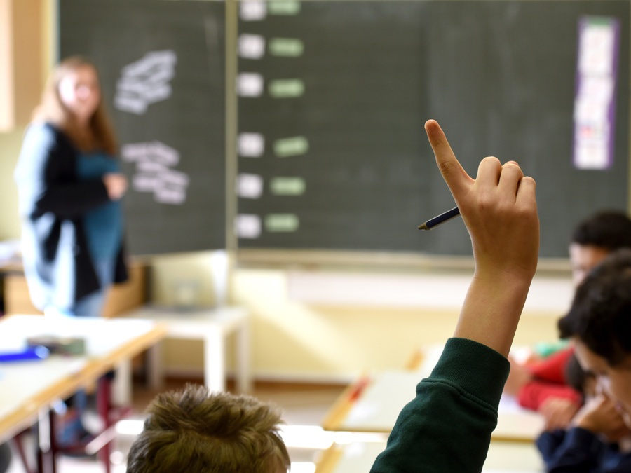Kind in einem Klassenzimmer, das die Hand hebt und den Finger streckt. Man sieht es von hinten.
