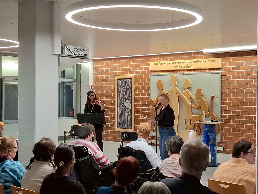 Das Publikum ist aufmerksam: Alle blicken nach vorn, zwei Personen sitzen im Rollstuhl. Im Hintergrund hängen ein Holzrelief stilisierter Menschen und ein Schild mit dem Spruch 'Wenn Du schnell gehen willst, gehe alleine. Doch wenn Du weit gehen willst, gehe mit anderen.