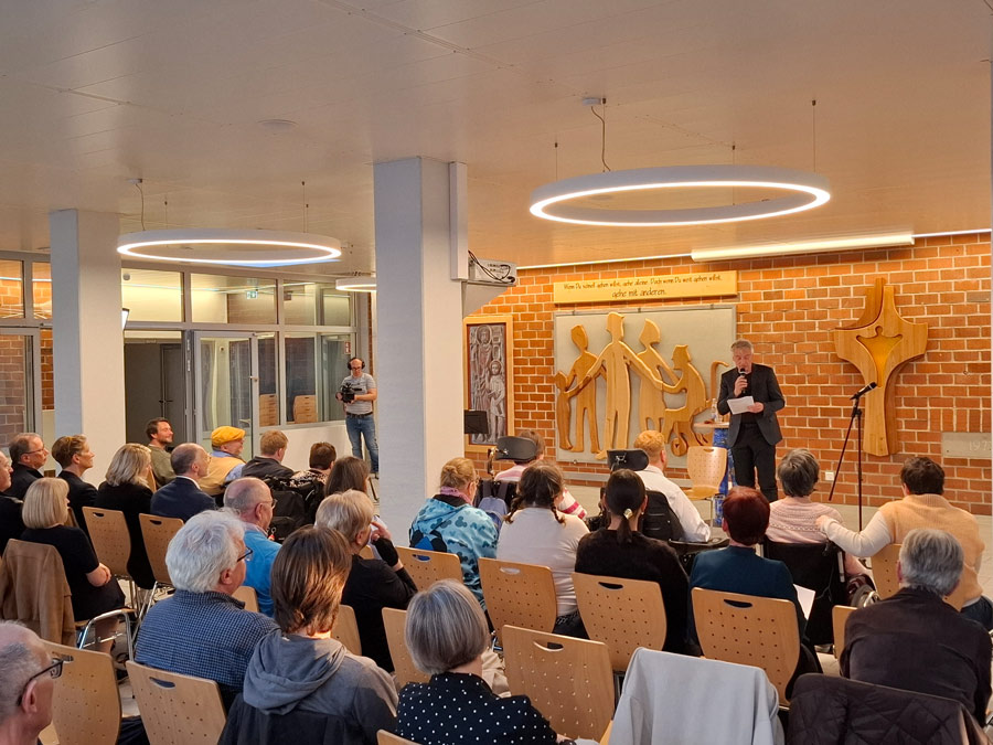 Prof. Dr. Christian Zürner steht im dunklen Anzug vor einer Backsteinwand und spricht in ein Mikrofon zu einem sitzenden Publikum, darunter auch Personen im Rollstuhl. An der Wand hängen ein Holzrelief stilisierter Menschen und ein Schild mit dem Spruch 'Wenn Du schnell gehen willst, gehe alleine. Doch wenn Du weit gehen willst, gehe mit anderen.' Im Hintergrund filmt ein Mann mit Kamera.