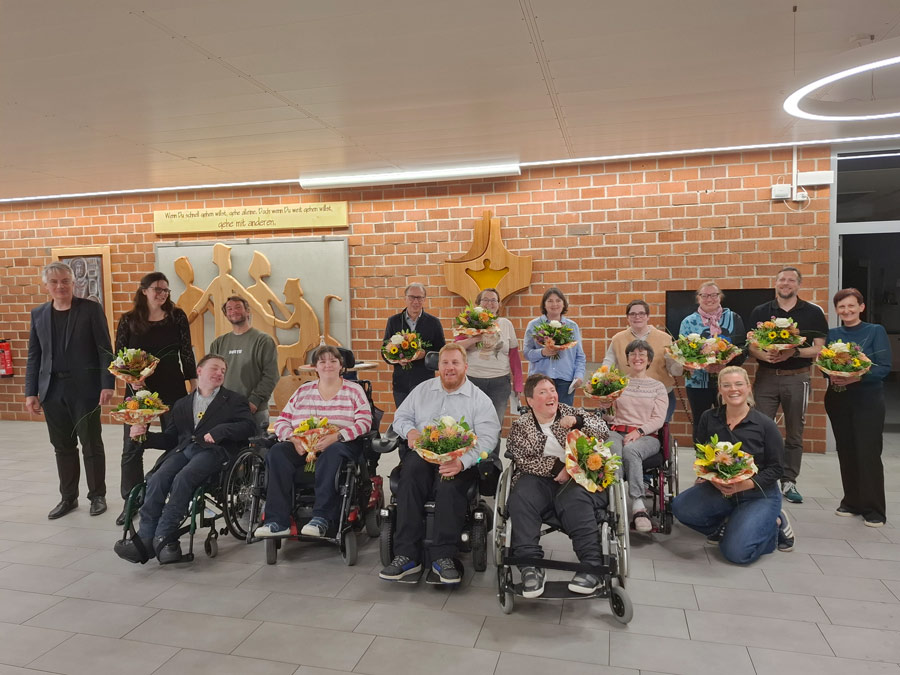 Gruppenfoto von 16 Personen in einem Saal vor einer Backsteinwand. Fünf Personen sitzen im Rollstuhl, eine kniet, zehn stehen dahinter. 14 von ihnen halten einen Blumenstrauß. Die Gruppe wirkt fröhlich: fast alle blicken lächelnd in die Kamera. An der Wand steht auf einem Schild: "Wenn Du schnell gehen willst, gehe alleine. Doch wenn Du weit gehen willst, gehe mit anderen